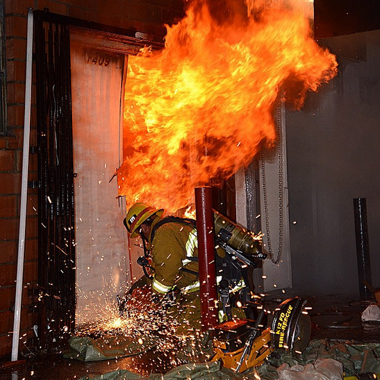A firefighter crouched using a rotary saw to force entry into a burning structure.